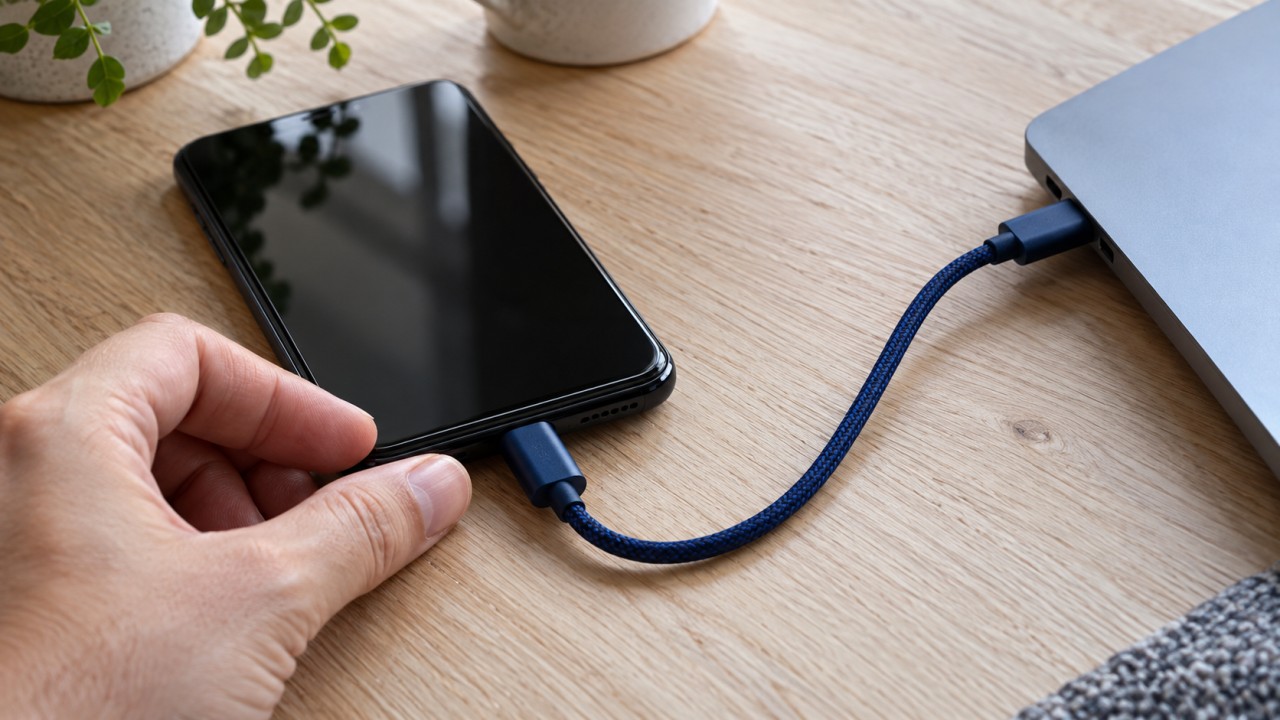 Celular conectado por cabo USB azul a um computador sobre uma mesa de madeira.
